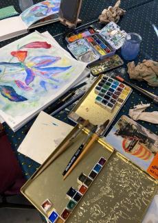 photo d'une table avec une aquarelle en cours, des pigments, pinceaux, palettes...